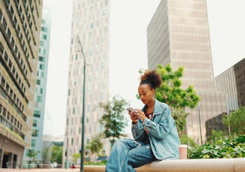 African girl with ponytail wearing denim jacket leaving voice message on mobile phone against modern city background.